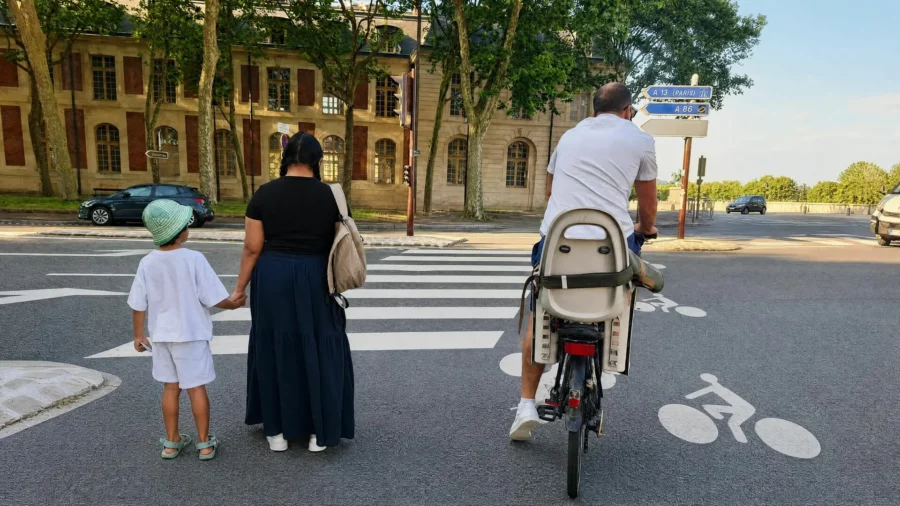 Une femme tenant la main d'un petit garçon et un homme à vélo avec un siège bébé attendent à un passage piéton devant un bâtiment historique à Paris par une journée ensoleillée.