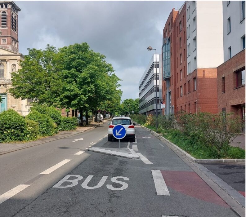 Image photoréaliste d'une rue urbaine à Roubaix, près de l'église et de bâtiments modernes. La chaussée est propre avec un asphalte neuf et des marquages blancs clairs, incluant l'inscription "BUS" et des flèches de direction. La végétation dense et verdoyante borde les trottoirs, créant un environnement écologique. Une voiture blanche est garée sur le côté droit, près d'un panneau de signalisation bleu. L'ensemble reflète un aménagement de qualité favorisant la mobilité douce et l'accessibilité PMR.